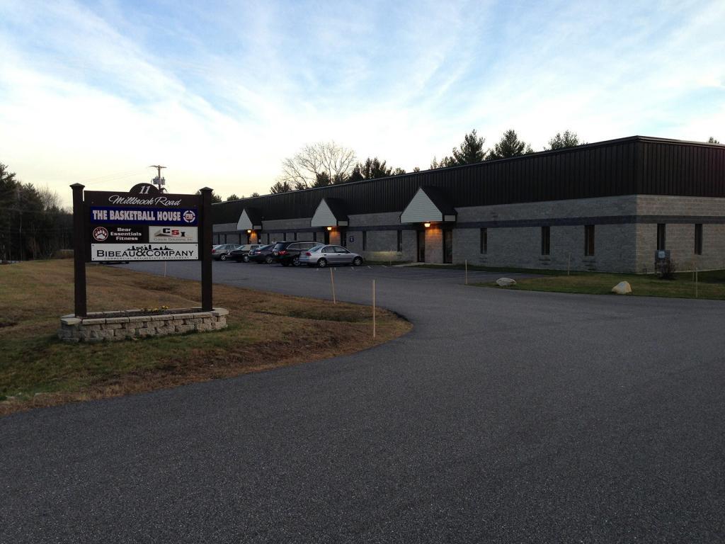 A long, one-story building with a large parking lot and several cars parked. A wooden sign in the foreground reads "The Basketball House" along with various business logos. The sky is clear with a hint of sunset.