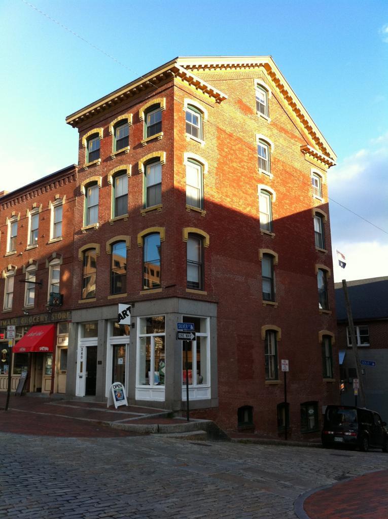 A historic three-story red brick building on a cobblestone street corner. The ground floor has shop windows and signs. The sky is clear with a few clouds, and the sun casts shadows on the building.