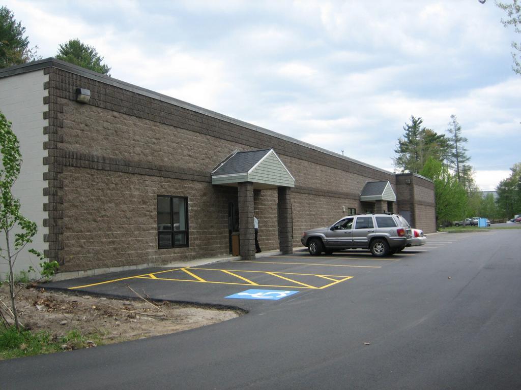 A gray SUV is parked in front of a one-story commercial brick building with two entrances, each with a small overhang. The parking lot includes a marked accessible parking space. Trees and overcast sky can be seen in the background.