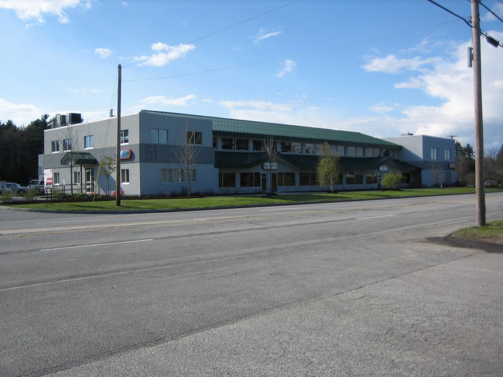 A wide view of a two-story commercial building with a green roof and multiple windows. It sits along a road with a grass lawn in front. The sky is partly cloudy, and there are trees in the background.