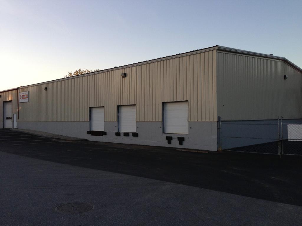 A large industrial warehouse with a beige metal exterior and three loading dock doors. The building is situated on an asphalt lot with a chain-link fence on the right side. The sky is clear, indicating a sunny day.