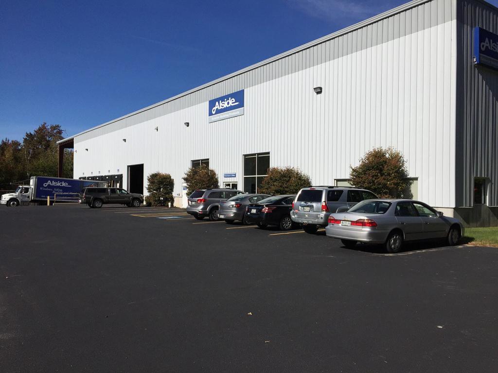 A commercial warehouse building with white metal siding and several cars parked in front. A sign on the building shows the name "Alside." Trees and a clear blue sky are visible in the background.