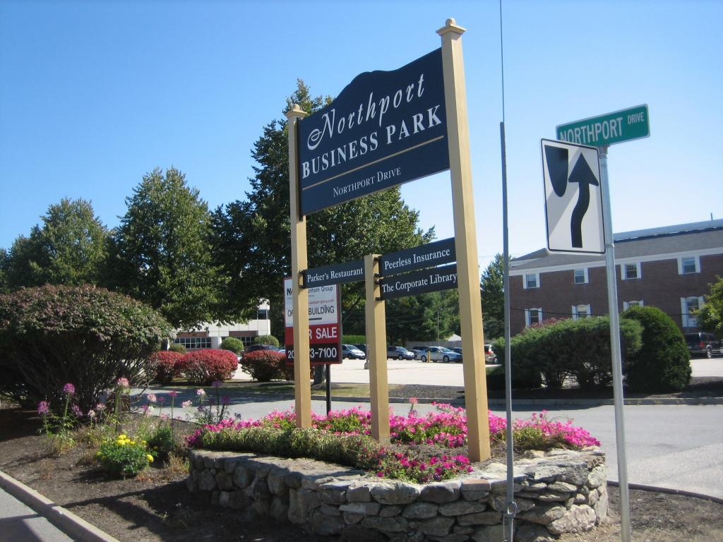 Sign at Northport Business Park entrance surrounded by flowers and shrubs. The sign lists nearby businesses, with a "for sale" sign below. A street sign for Northport Drive is visible against a clear blue sky. Parking lot and building in the background.