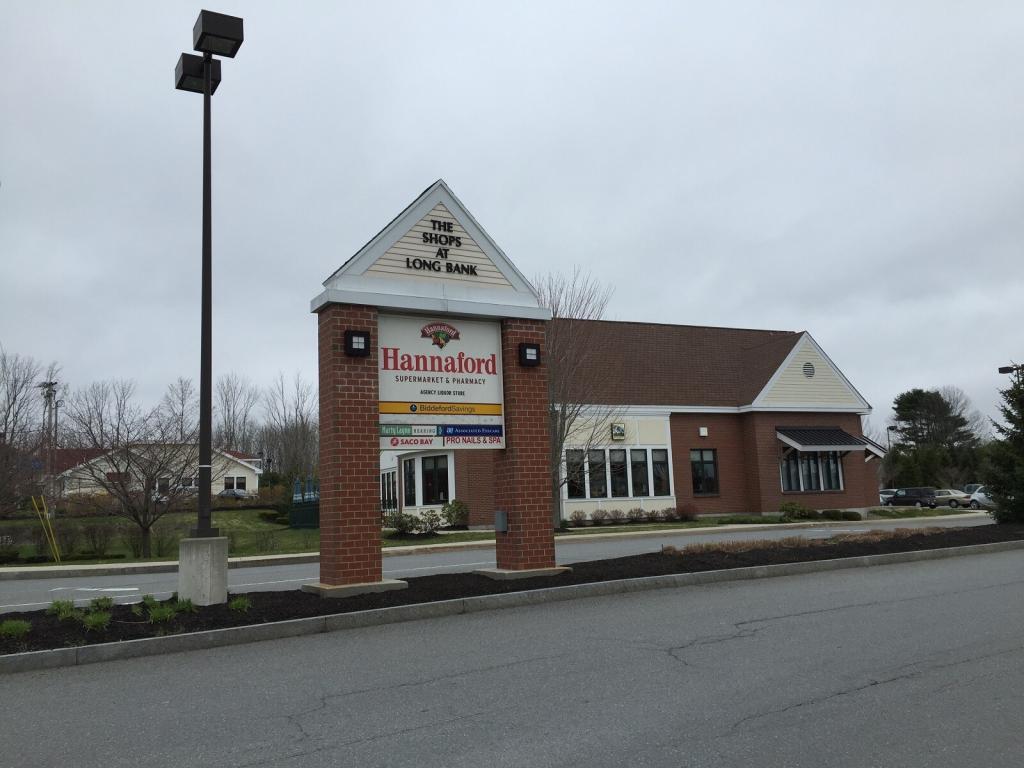 A shopping plaza with a sign reading "Hannaford Supermarket & Pharmacy" in front of a brick building. The sky is overcast, and the area is surrounded by trees and a parking lot.