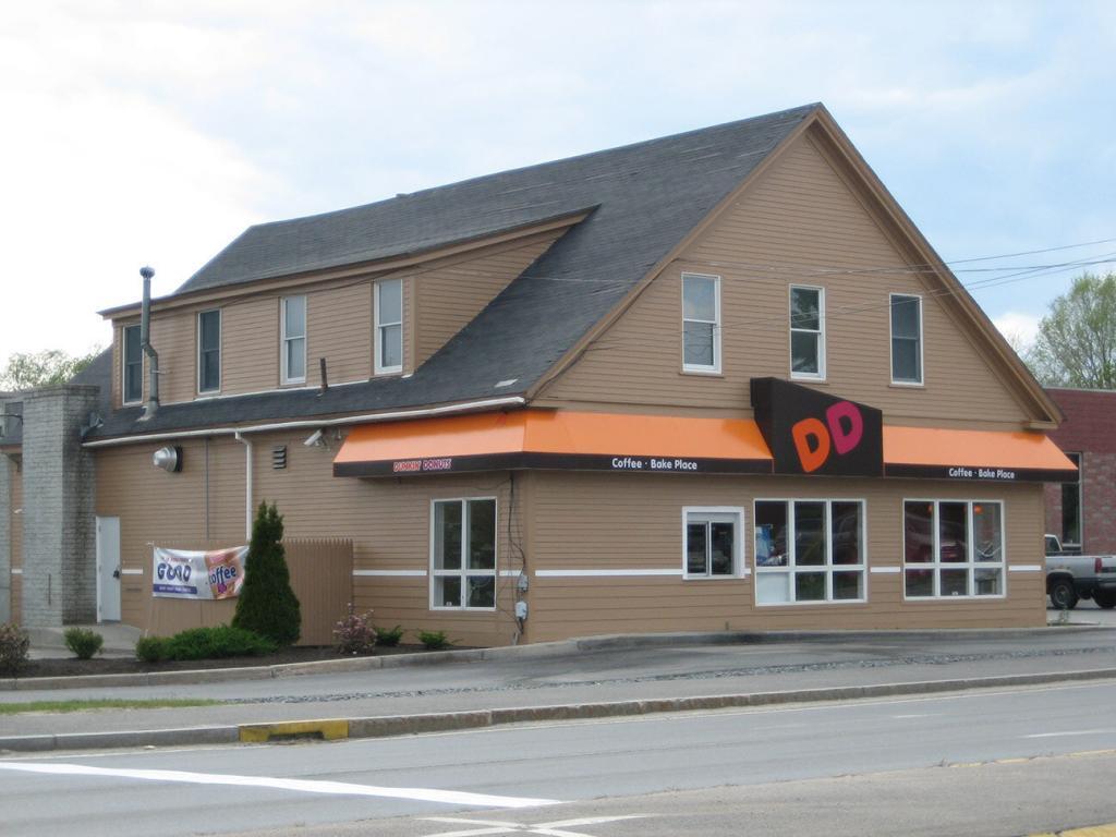 A standalone Dunkin' Donuts store with a beige exterior and typical orange and pink signage. The building has multiple windows and a sloped roof. It stands along a roadway with visible street markings.