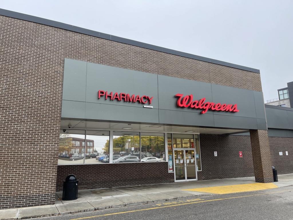 A Walgreens store exterior with brick walls and a grey canopy displaying the red signs "Pharmacy" and "Walgreens." The entrance has glass doors and windows.