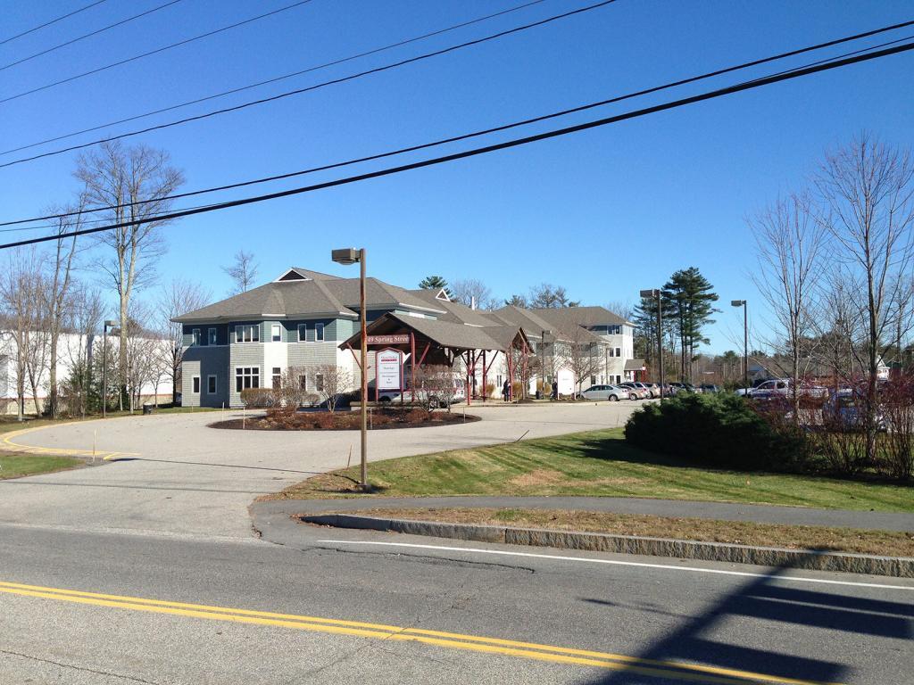 A large, two-story building with a gray roof and red wooden supports at the entrance. It's surrounded by a parking lot, trees, and a clear blue sky. A road with yellow lines is in the foreground, along with power lines overhead.