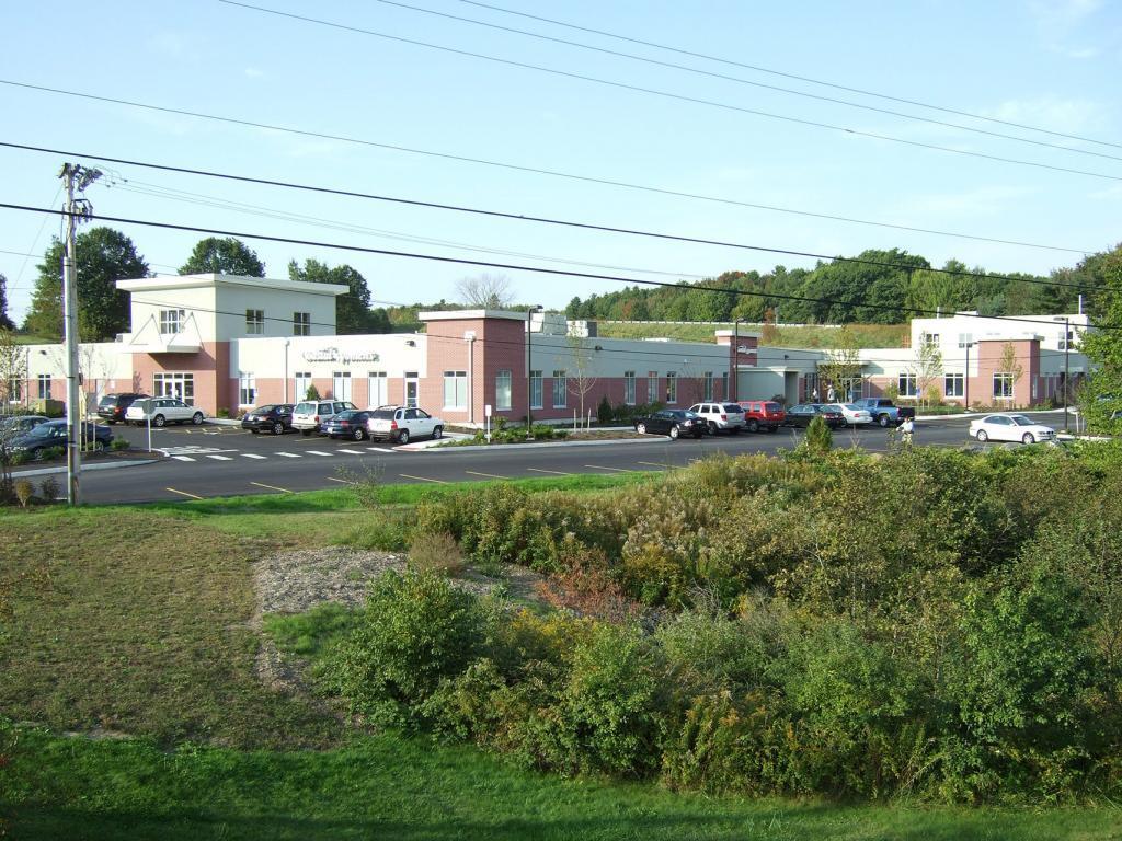 A single-story, modern building with a red and white facade, surrounded by a parking lot with several cars. There is greenery in the foreground and trees in the background under a clear sky.