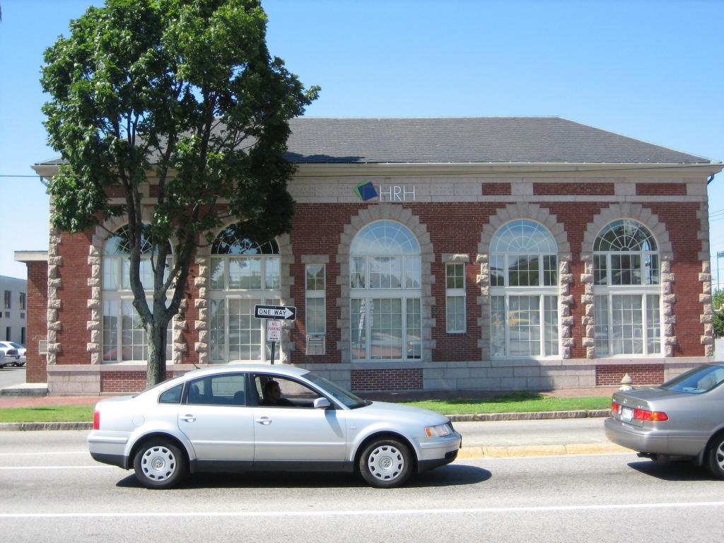 A red brick building with large arched windows and a sign reading "HRH" is positioned near a street. A silver car is driving by in the foreground, and a large green tree is beside the building.