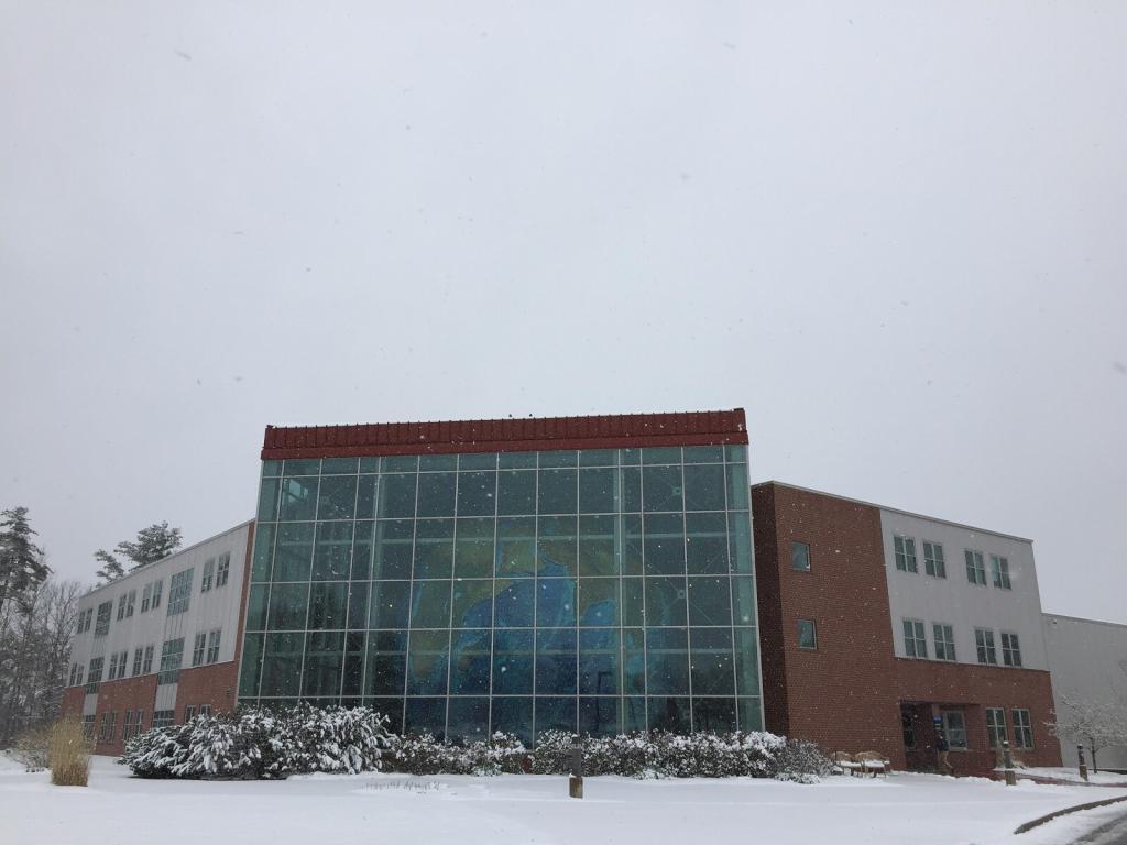 A modern building with large glass windows and brick accents stands surrounded by snow-covered ground and bushes. The sky is overcast, and snow is lightly falling, creating a serene winter scene.