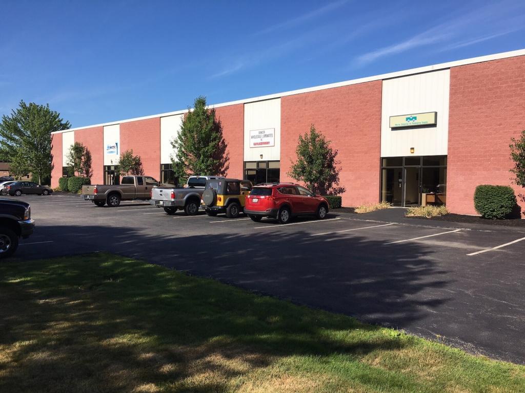 A row of single-story commercial buildings with red brick facades. Several vehicles are parked in front, and small trees line the sidewalk. The sky is clear and blue.
