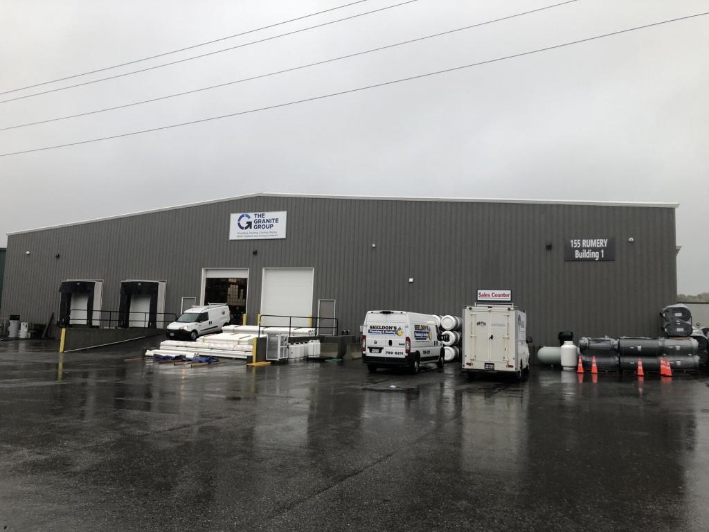 A warehouse building with a gray exterior on a rainy day. Several vans are parked outside, with some loading dock doors open. Stacks of materials are visible near the entrance. The ground is wet, and traffic cones are placed nearby.