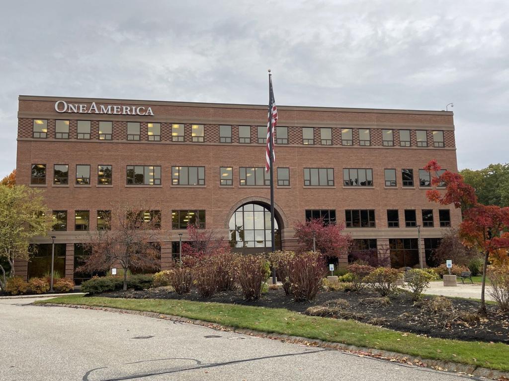 A red brick office building with "OneAmerica" on top. The building has four stories and a large glass archway entrance. An American flag is in front, surrounded by landscaped shrubs and trees with autumn foliage. Overcast sky above.
