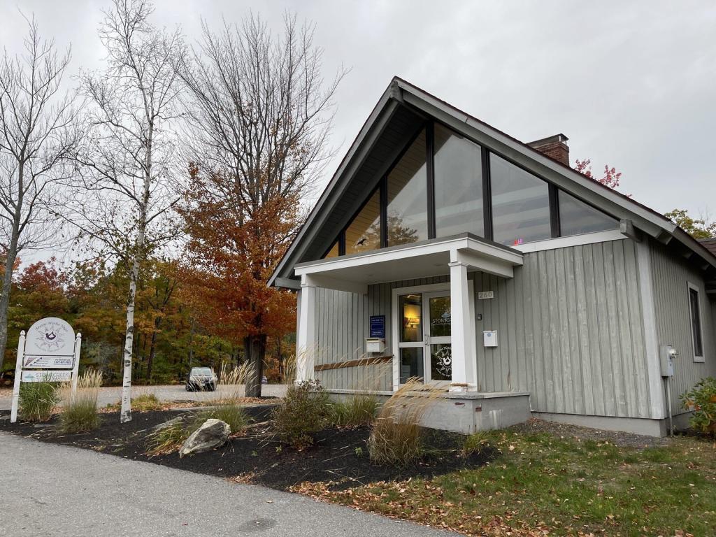 A small, modern building with a steep pitched roof and large front windows, surrounded by autumn trees with red and orange leaves. A white sign is positioned near the entrance, and a driveway is visible to the left.