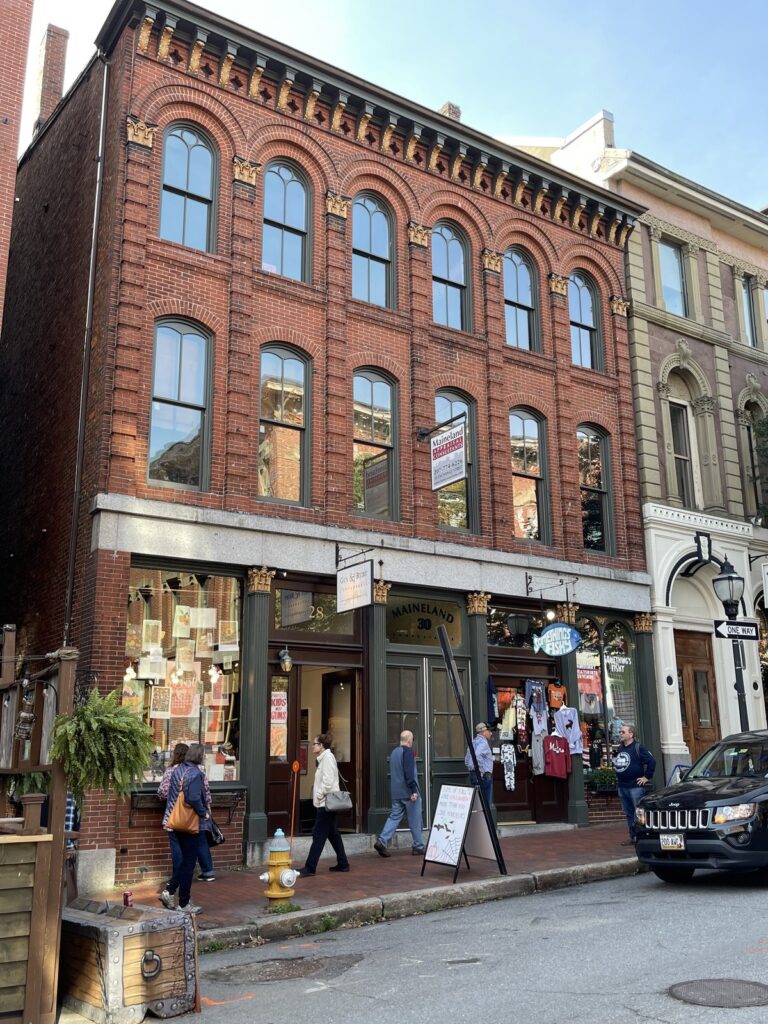 A street view of a brick building with arched windows. The ground floor has businesses with signs and people walking past. There's a parked black car in front and a sandwich board on the sidewalk.