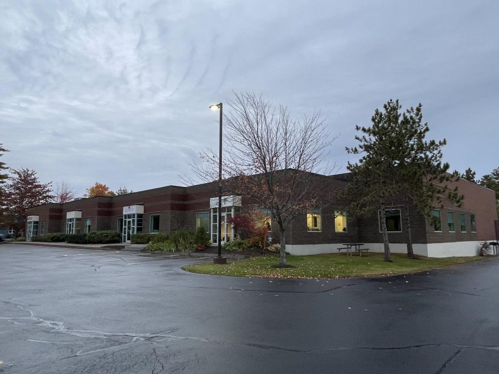 A large brick building with multiple doorways and windows, set against a cloudy sky. Leafless trees and a few evergreens surround the area. A single streetlamp illuminates the wet asphalt parking lot in the foreground.