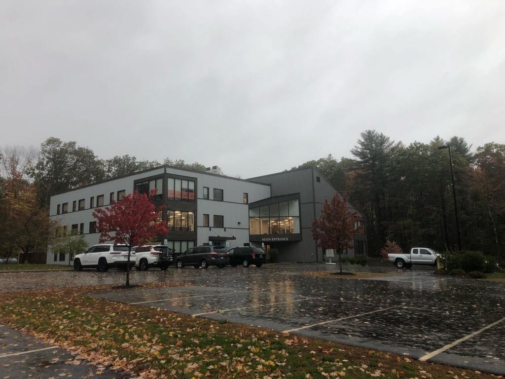 A modern, multi-story building with large windows sits in a parking lot. Several vehicles are parked in front. The ground is wet from rain, and there are autumn leaves scattered around with trees in the background.