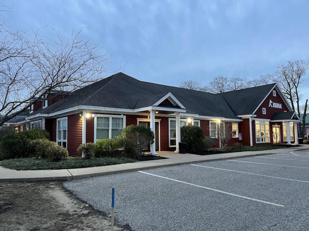 A single-story red building with a gray roof stands in the early evening light, under the care of Bibeau Property Management. The entrance is flanked by shrubs and a small ramp. A sign reads "KUMON" on the building. The parking lot in front is empty, and bare trees surround the area.