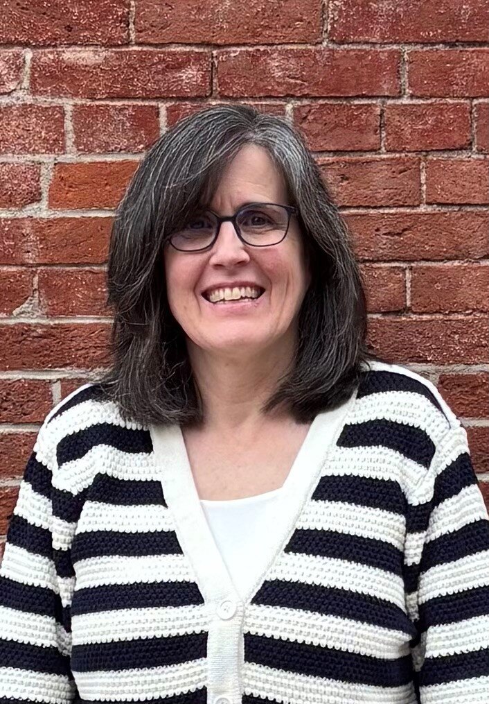 A photo of Bibeau staff member Deb Young, showing a woman in front of a brick wall wearing a black and white sweater.